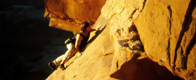 Tom Cruise escaladant une paroi rocheuse à mains nues dans une scène du film Mission : Impossible 2.